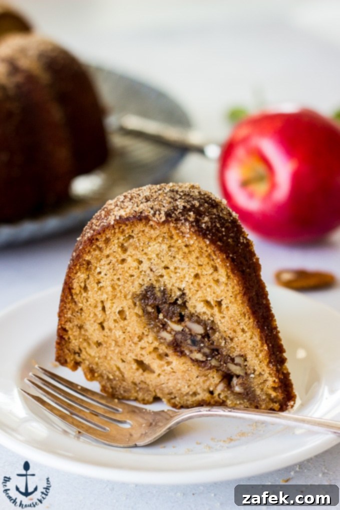 Slice of Apple Cider Doughnut Bundt Cake with Pecan Brown Sugar Ripple on a white plate with a silver fork and an apple in the background.