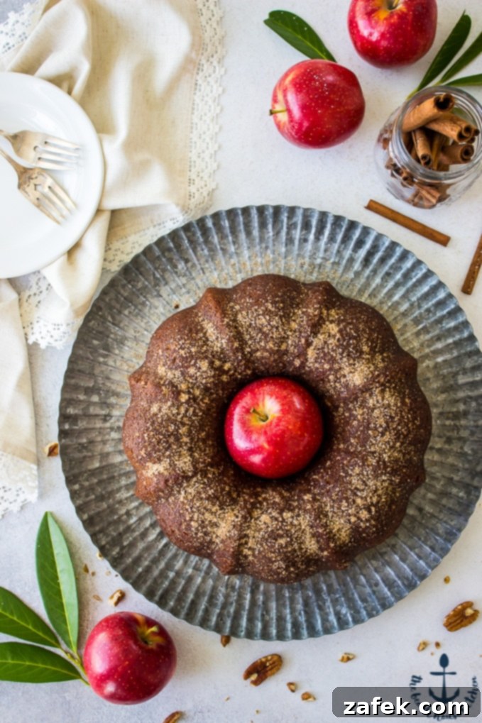 Overhead photo of Apple Cider Doughnut Bundt Cake with Pecan Brown Sugar Ripple with a red apple in middle of cake on a silver tray surrounded by red apples and cinnamon sticks.