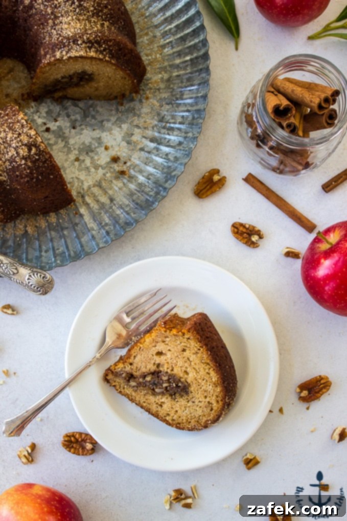 A slice Apple Cider Doughnut Bundt Cake with Pecan Brown Sugar Ripple on a white plate with remainder of bundt cake on silver tray in background.