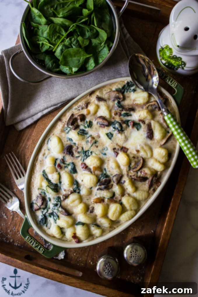 An oval dish filled with rich Mushroom Florentine Gnocchi, garnished with fresh herbs, resting on a wooden tray beside a colander brimming with vibrant green spinach leaves.