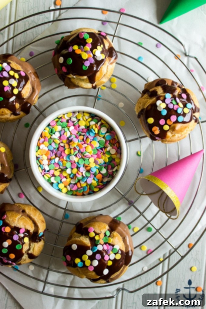 Nutella-Cream-Puffs Overhead photo of Nutella Cream Puffs drizzled with chocolate and sprinkles on circular silver cooling rack with white bowl of sprinkles in center with mini party hats.