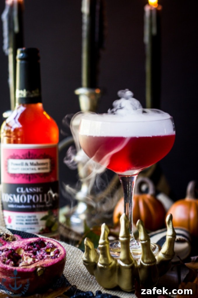 Vampires-Venom-Cocktail Red Vampire's Venom Cocktail with dry ice smoke bubbling in a gold skeleton hand with candles and cocktail bottle in background.