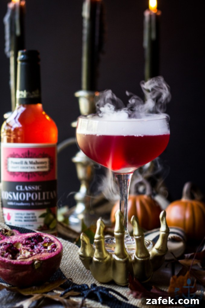 Vampires-Venom-Cocktail Red Vampire's Venom Cocktail with dry ice smoke bubbling in a gold skeleton hand with candles and cocktail bottle in background.