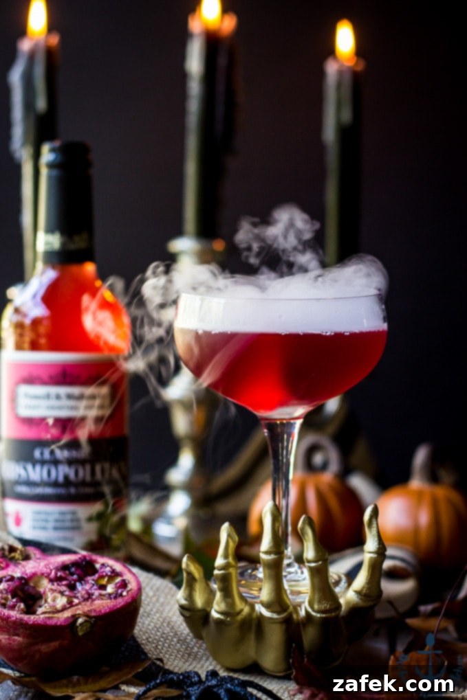 Vampires-Venom-Cocktail Red Vampire's Venom Cocktail with dry ice smoke bubbling in a gold skeleton hand with candles and cocktail bottle in background.