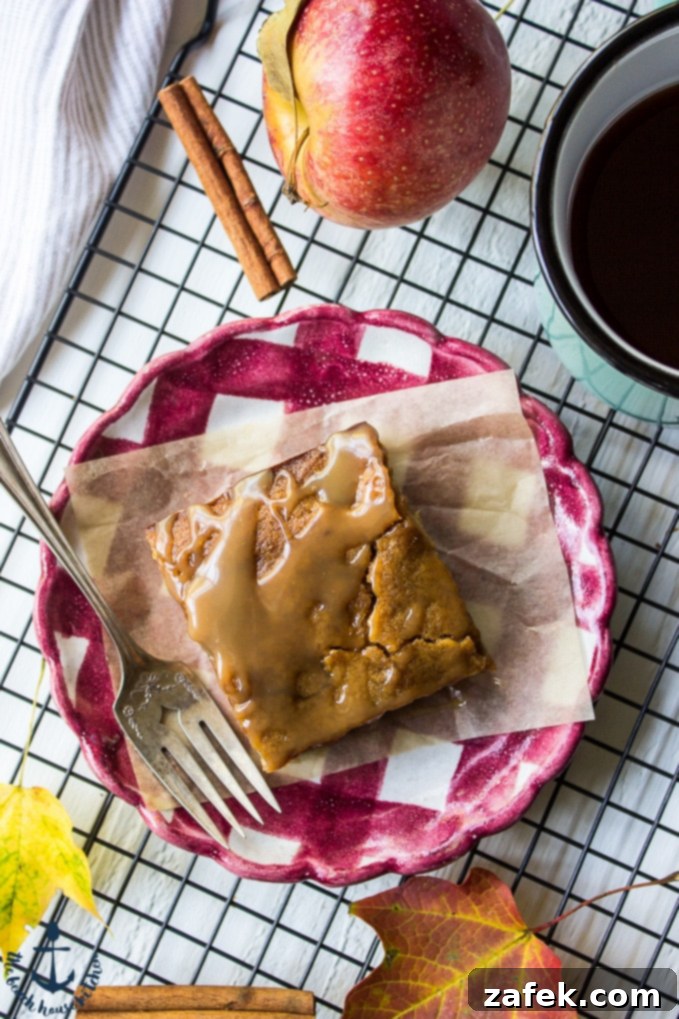 A close-up of a Caramel Glazed Apple Butter Blondie on a red and white check plate, with a fork, a fresh apple, and scattered autumn leaves in the background, emphasizing the seasonal delight.