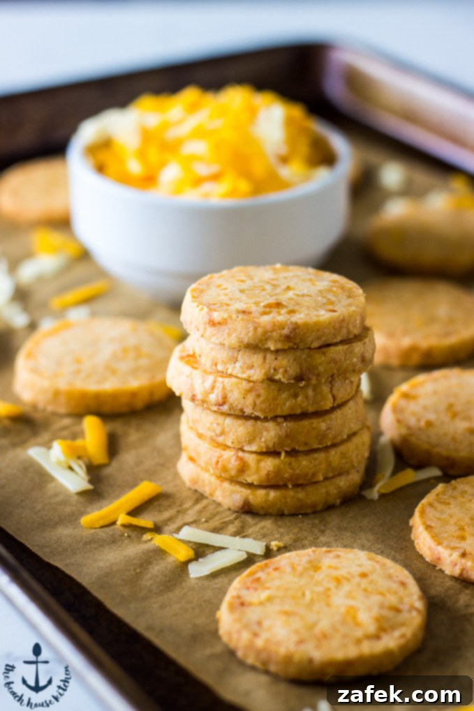 Stack of golden-brown Cheddar Cheese Coins artfully arranged on a baking sheet lined with parchment paper, with a pristine white bowl brimming with freshly shredded cheddar cheese in the background. The image evokes a sense of homemade goodness and rich flavor, highlighting the key ingredient and the crisp texture of the finished product.