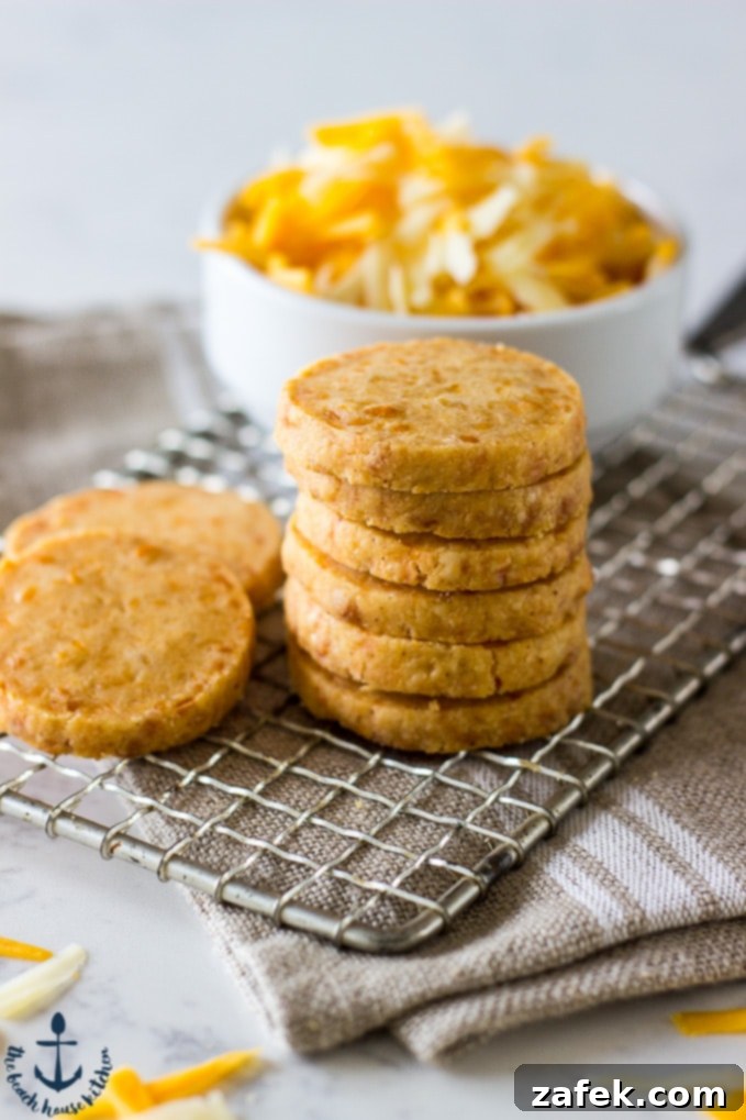 A stack of perfectly golden-brown Cheddar Cheese Coins cooling on a wire rack, with a blurred background showing a white bowl of shredded cheddar, suggesting the delicious journey from raw ingredient to irresistible snack. The image conveys crispness and fresh baking.