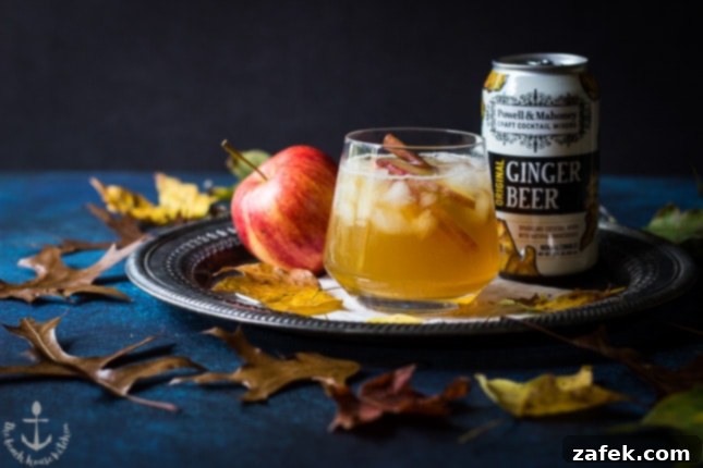 Fall-Dark-Stormy The Fall Dark 'N' Stormy cocktail on a silver tray with an apple and a can of ginger beer, surrounded by vibrant fall leaves, creating a festive scene.