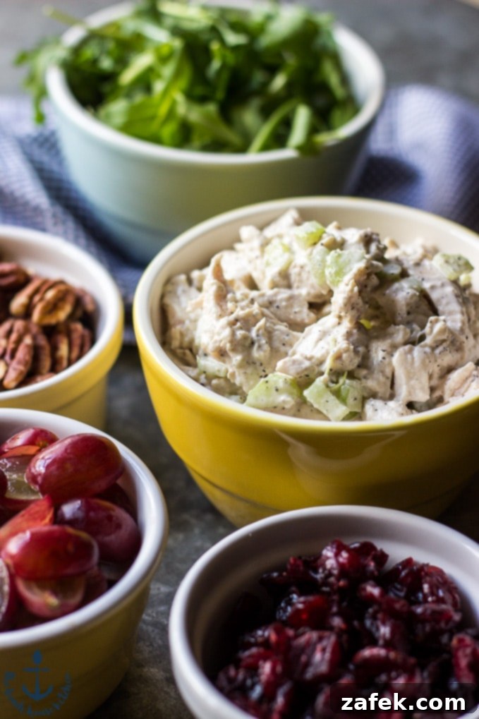 A close-up shot of the vibrant components of turkey salad: a yellow bowl of creamy turkey salad, with separate bowls of bright red cranberries, juicy red grapes, crunchy pecans, and fresh green arugula leaves.
