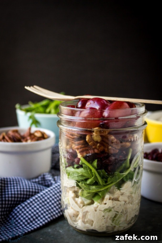 A perfectly layered Turkey Salad with Grapes, Pecans, and Cranberries in a Jar, with a fork resting on top, set against a blue and white checked napkin, ready for serving.