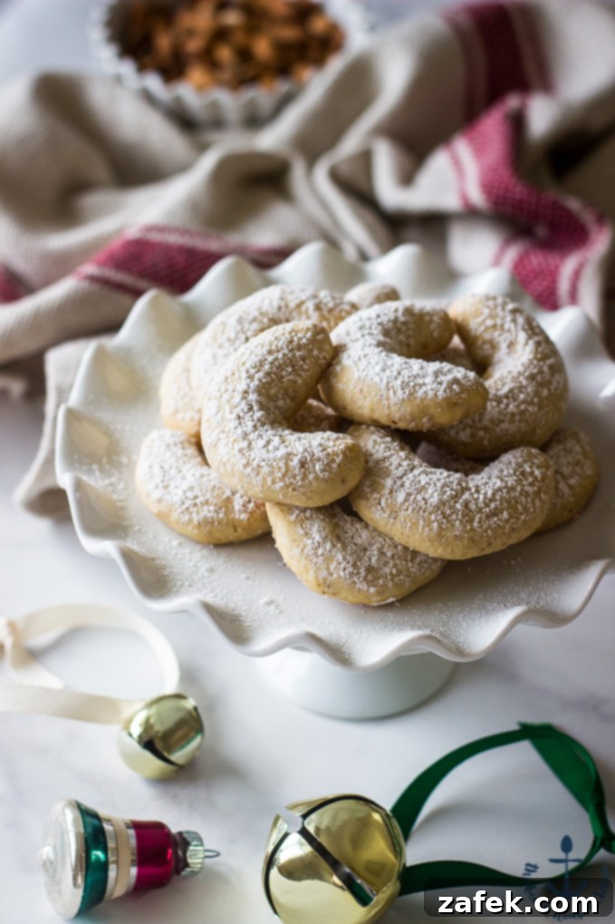 Almond Crescents dusted with confectioners' sugar on a white pedestal plate with festive jingle bells in the background, highlighting their elegant and simple holiday charm.