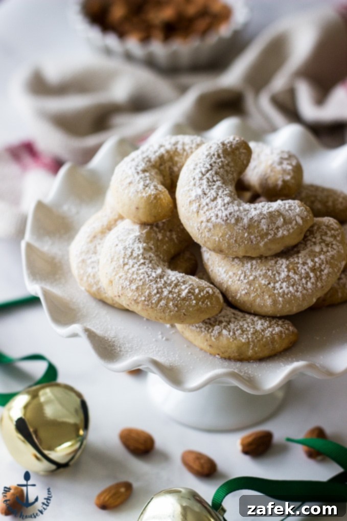 Another view of Almond Crescents beautifully dusted with confectioners' sugar, arranged on a white pedestal plate with sparkling jingle bells in the soft-focus background.