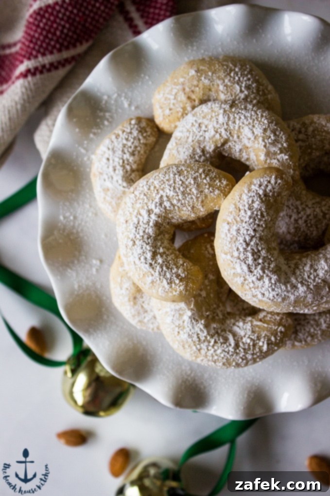 Almond Crescents artfully arranged on a pristine white plate, generously dusted with confectioners' sugar, with a subtle festive jingle bell decoration in the background.