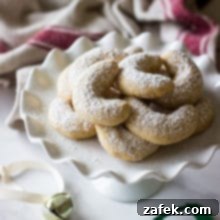 Almond Crescents dusted with confectioners' sugar on white pedestal plate with jingle bells in background.