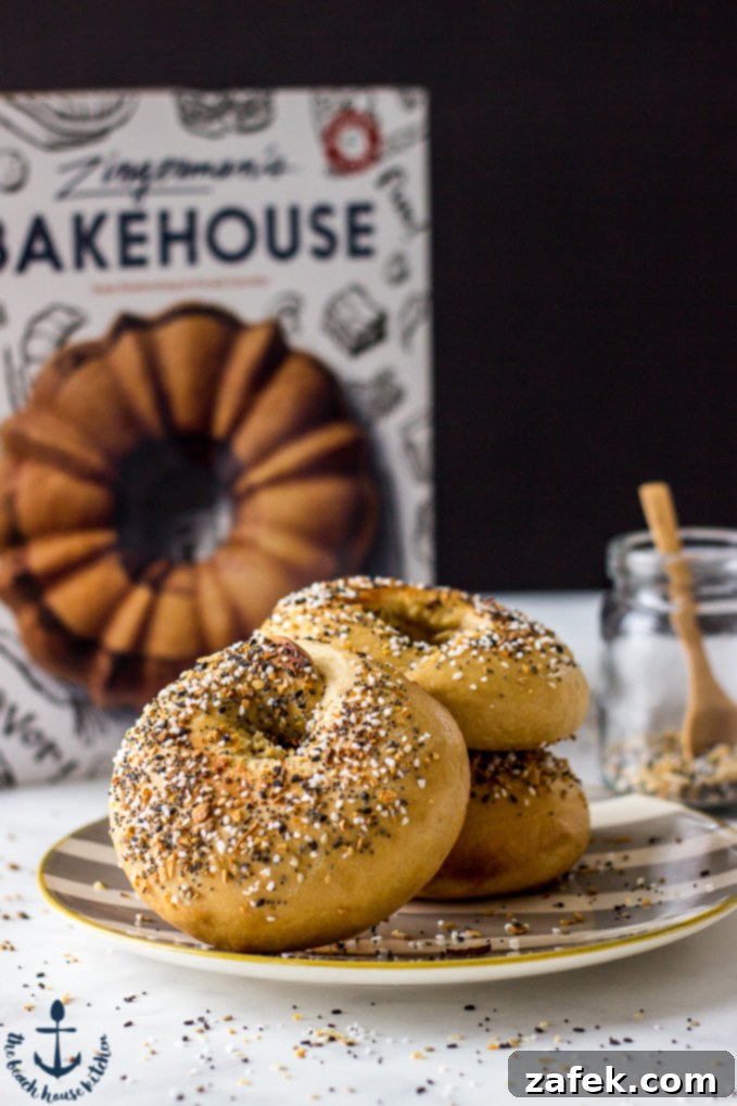 Homemade Everything Bagels Three golden-brown Everything Bagels stacked on a plate with a cookbook in the soft-focus background.