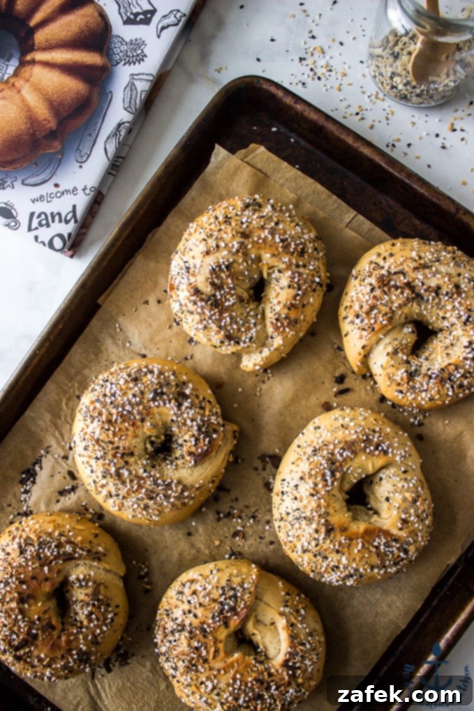 Easy Everything Bagel Recipe Freshly baked Everything Bagels cooling on a baking sheet lined with parchment paper, with a cookbook and seasoning jar nearby.