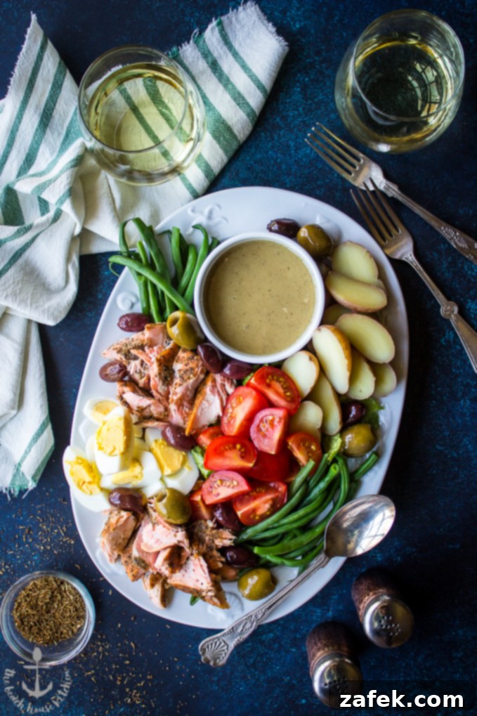 Overhead shot of Salmon Nicoise Salad with Dijon Vinaigrette on a blue board surrounded by wine glasses, salt and pepper shakers, forks and spices, highlighting fresh ingredients and vibrant colors.