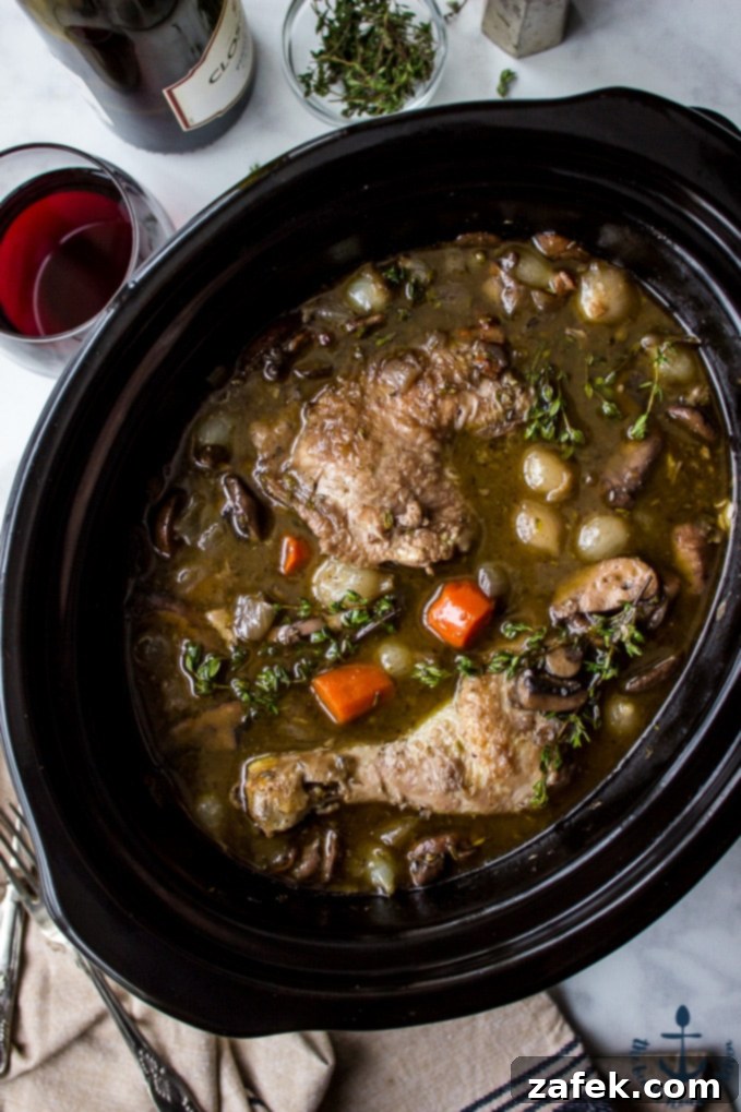 Slow Cooker Coq Au Vin Overhead photo of Slow Cooker Coq Au Vin in a black slow cooker with a bottle of wine , glass of wine and salt and pepper shakers in background.