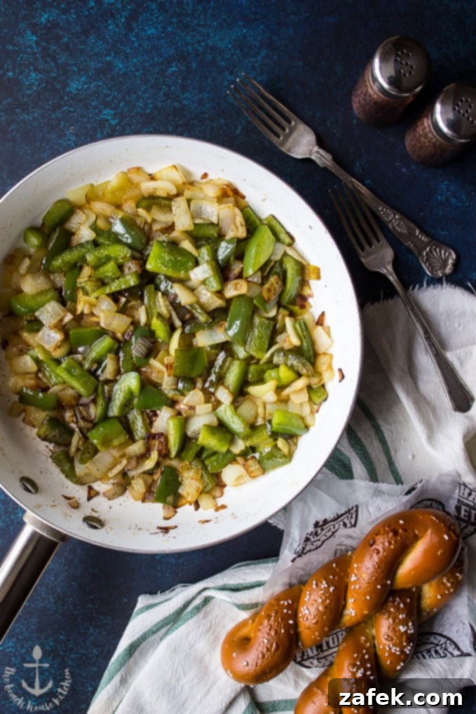 A close-up shot of green bell peppers and onions sautéing in a skillet, with two soft pretzels in the blurred background, capturing an essential step in making the dish.