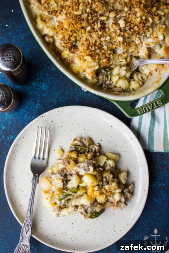 A white plate featuring a serving of Philly Cheesesteak Macaroni and Cheese with a silver fork, next to a larger oval baking dish brimming with the same delicious pasta, ready to be enjoyed.