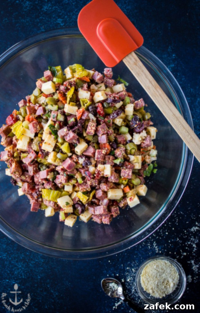 Overhead photo of Muffuletta Dip in a glass bowl with an orange rubber spatula resting on top.