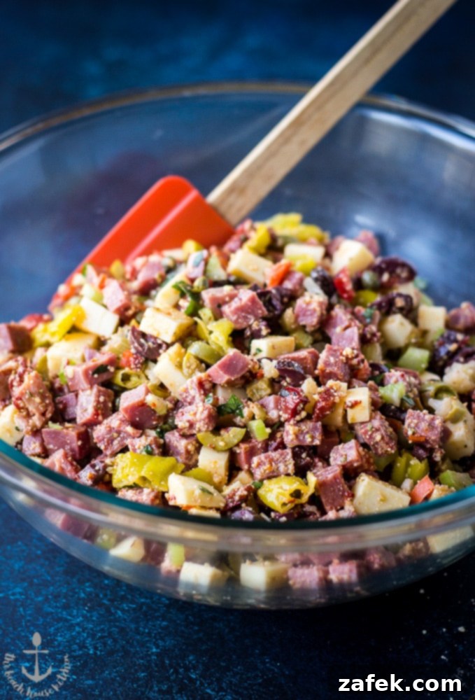 Muffuletta Dip in a glass bowl with a rubber spatula on a blue background.