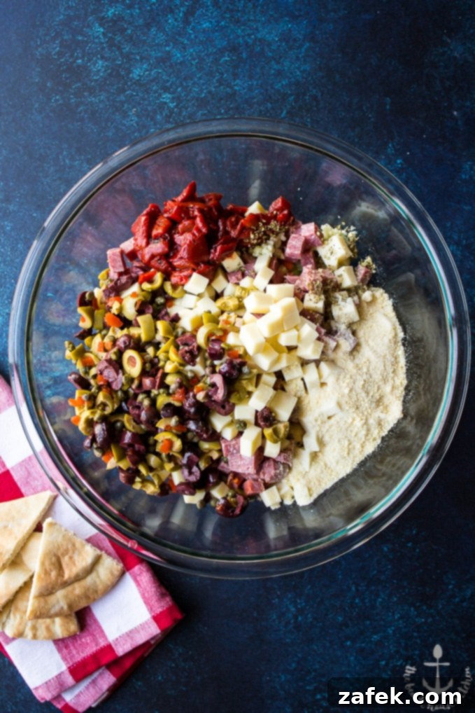 Overhead photo of Muffuletta Dip ingredients in a glass bowl on a blue background with a red and white checked napkin.