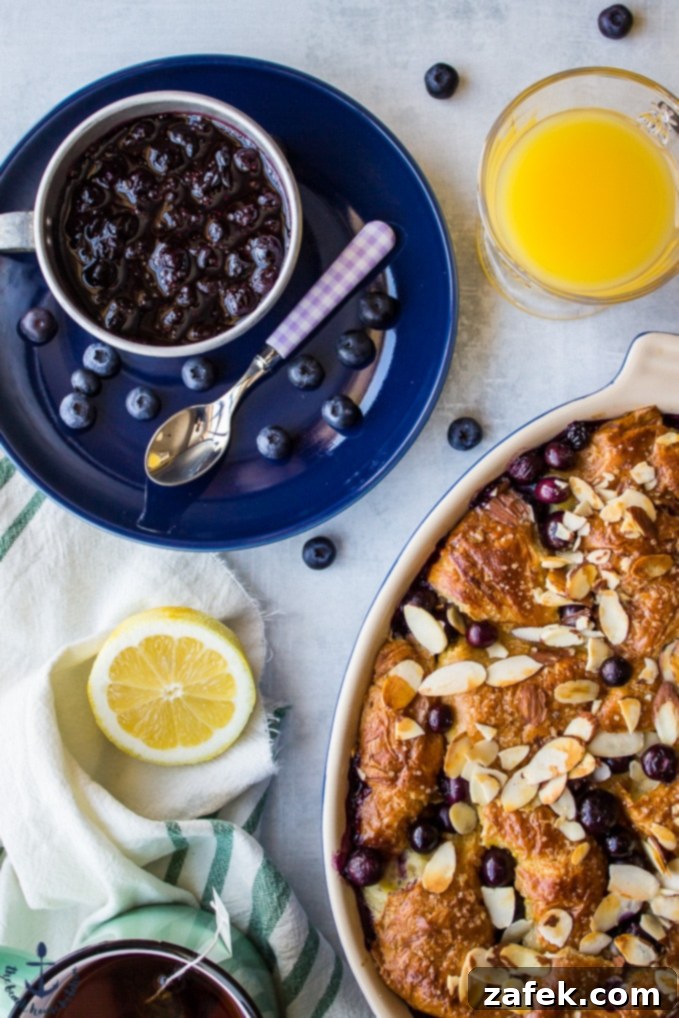Blueberry Croissant Bread Pudding with Blueberry Sauce Overhead shot of Blueberry Croissant Bread Pudding in a ceramic dish, accompanied by a small bowl of blueberry sauce on a blue plate.