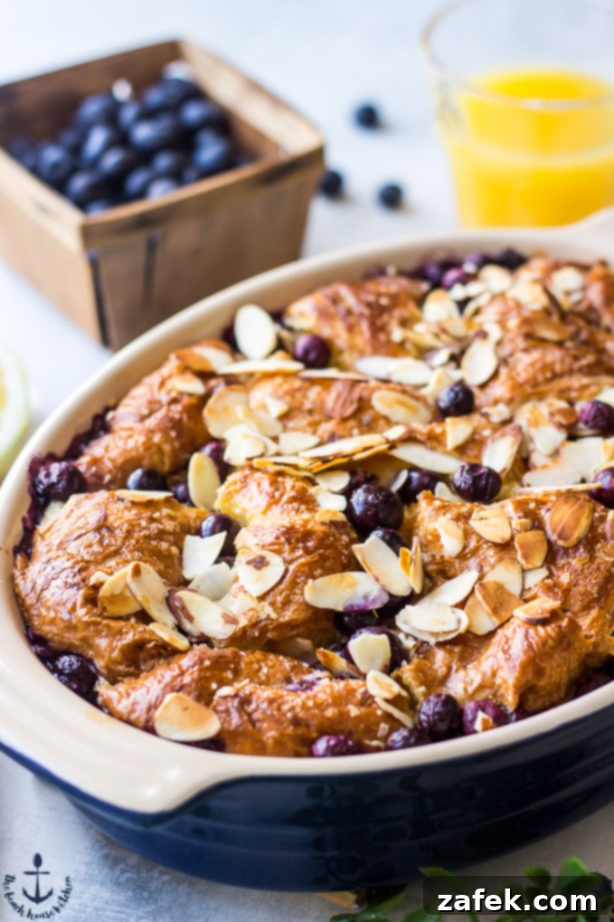 Blueberry Croissant Bread Pudding with Blueberry Sauce A serving of Blueberry Croissant Bread Pudding in a blue oval dish, with fresh blueberries and a glass of orange juice blurred in the background, hinting at a delightful breakfast.