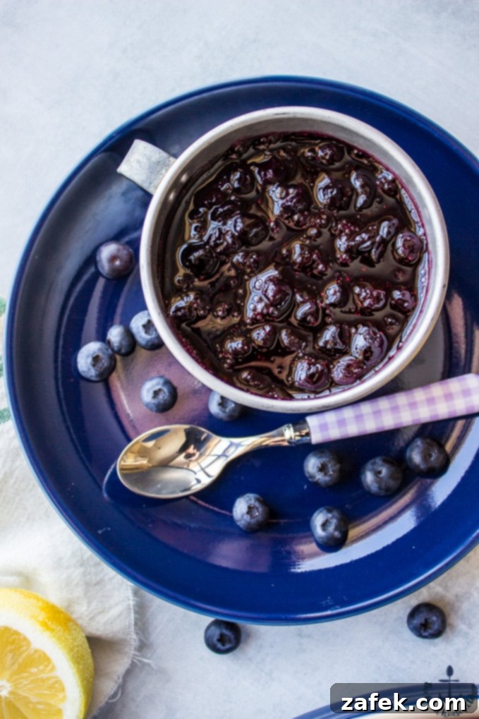 Blueberry Croissant Bread Pudding with Blueberry Sauce Close-up overhead shot of homemade blueberry sauce in a small silver cup, resting on a blue plate with a spoon, highlighting its rich texture.