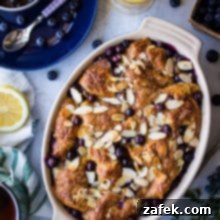 Blueberry Croissant Bake with Berry Compote 2 Overhead photo of Blueberry Croissant Bread Pudding in an oval dish with a cup of blueberry sauce on a blue plate, a glass of orange juice and a half of a lemon.