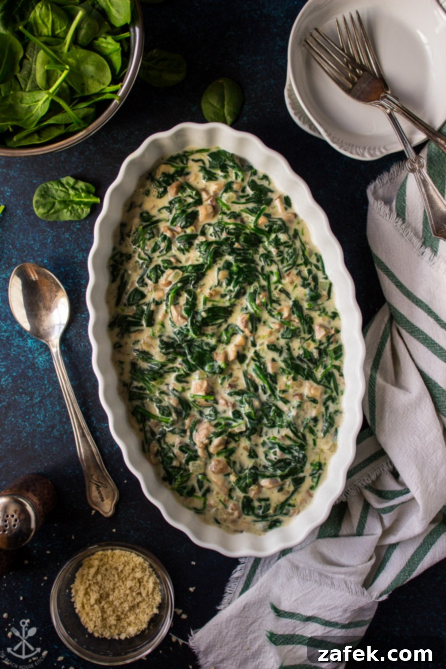 Overhead photo of an oval white dish filled with creamed spinach and mushrooms on a rustic blue background with a serving spoon off to the side