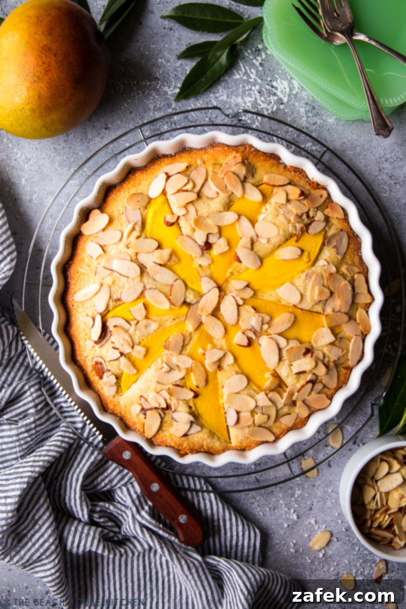 Overhead photo of a mango cake in a round white scalloped baking dish, garnished with fresh mango slices and toasted almonds.