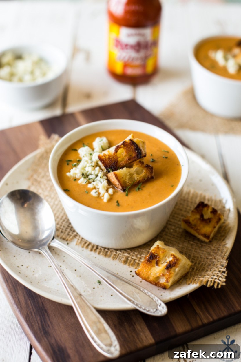 A bowl of buffalo cauliflower soup on a white plate with two silver spoons resting beside it, garnished with blue cheese crumbles and croutons