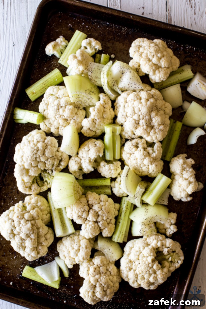A baking sheet filled with raw cauliflower florets, sliced yellow onion, and chopped celery, drizzled with olive oil and ready for roasting