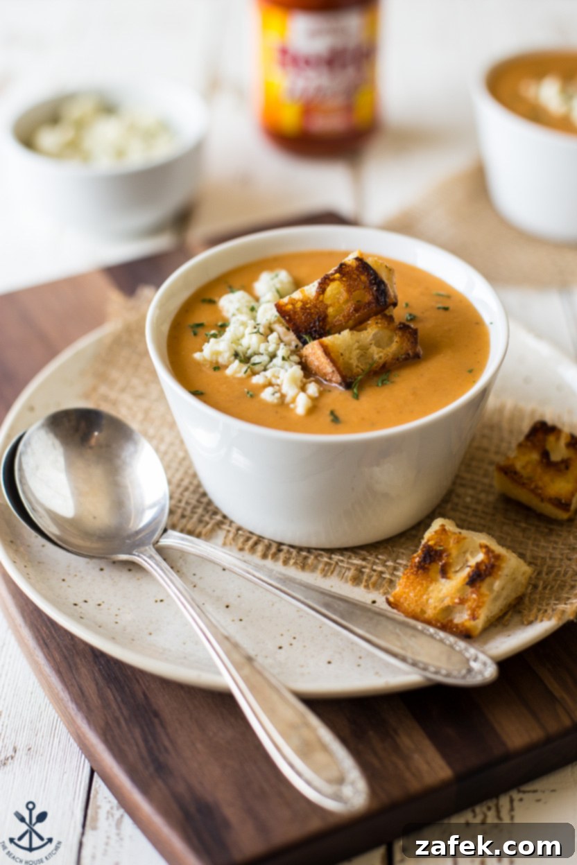 Up close photo of a bowl of creamy buffalo cauliflower soup on a white plate with two silver spoons, showcasing the texture and vibrant colors of the garnishes