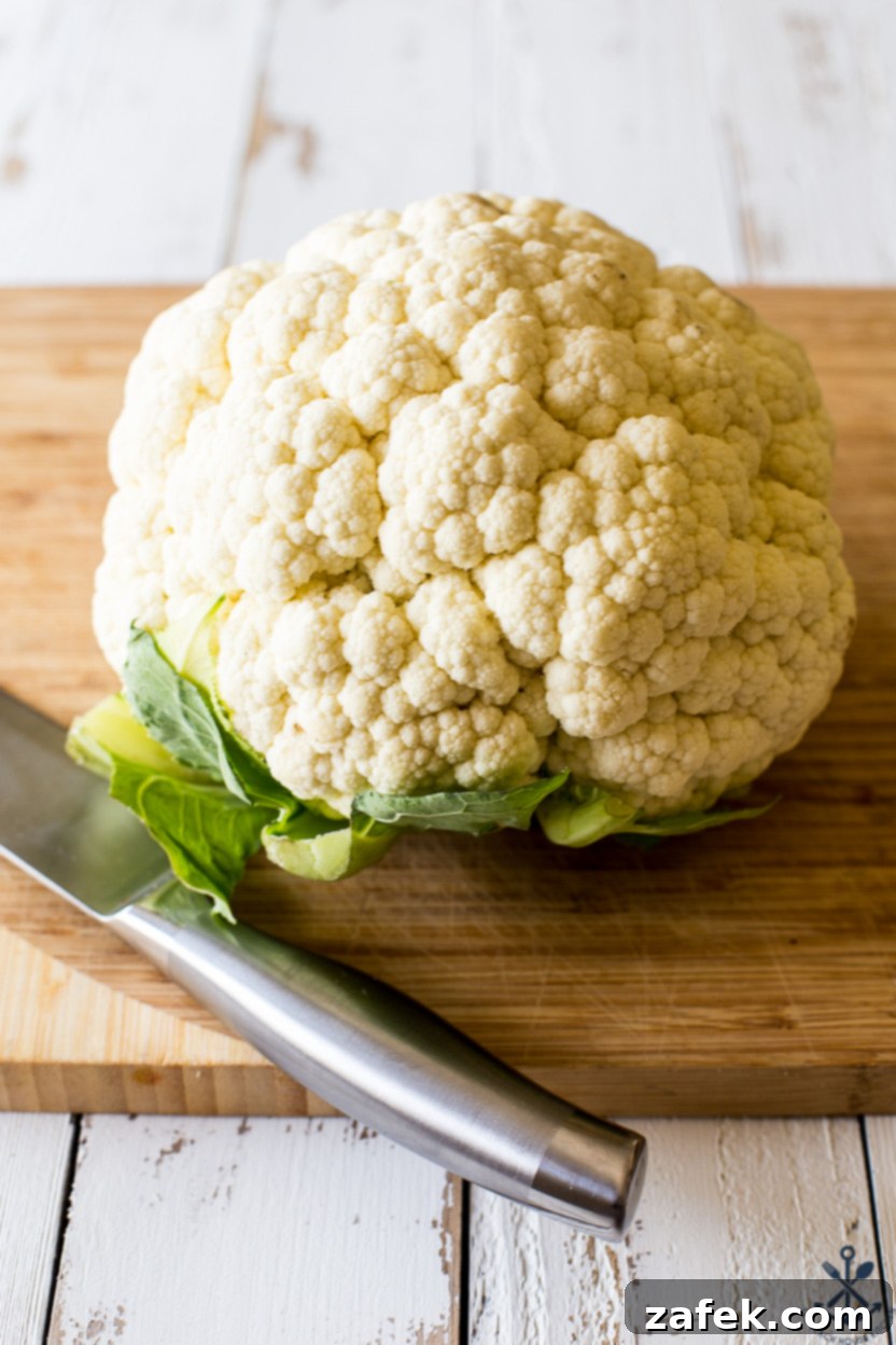 A whole head of fresh cauliflower resting on a wooden cutting board next to a large silver chef's knife