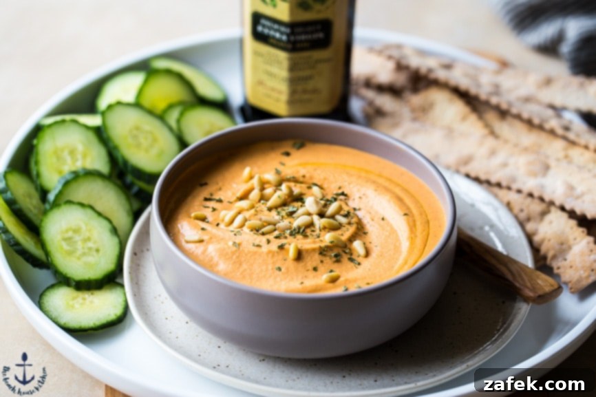 An overhead shot of a vibrant platter with White Bean Roasted Red Pepper Hummus, sliced cucumbers, flatbread crackers, and olives.