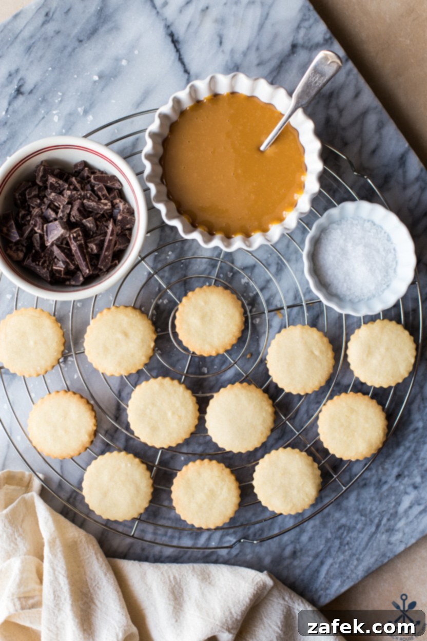 Copycat Twix Cookies 5 Overhead photo of cut out cookies on a round wire rack with a bowl of melted caramel, a bowl of chocolate pieces and a small bowl of salt