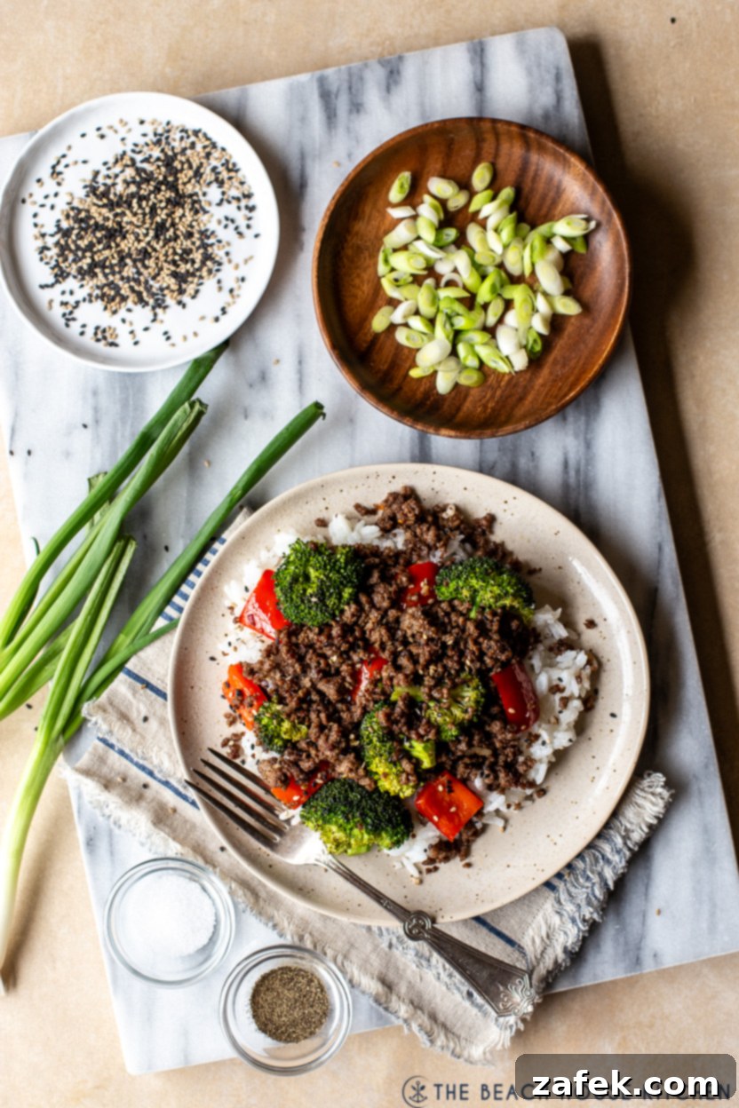 Overhead photo of a plate of Korean ground beef with broccoli and peppers on a marble board with a plate of sliced green onions and a plate of sesame seeds