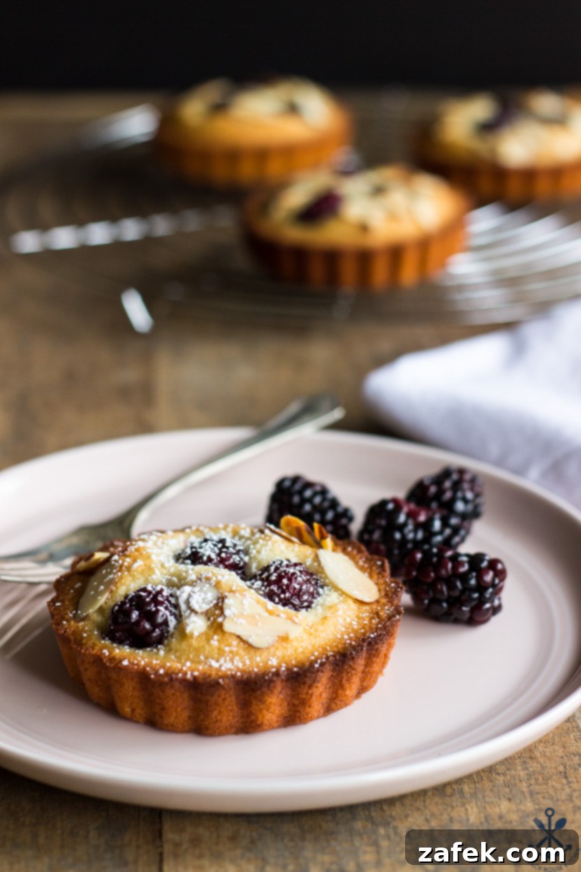 Elegant Blackberry Almond Bites 4 A perfectly baked financier on a plate with a fork and a scattering of fresh blackberries