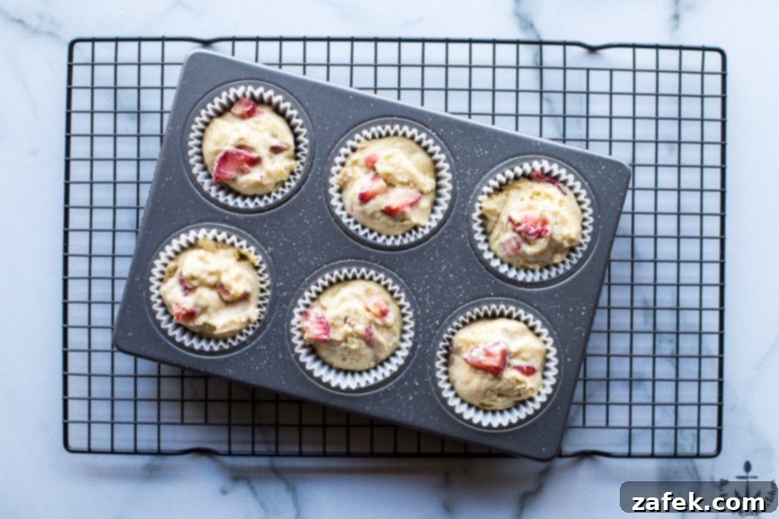 Sunshine Strawberry Lemon Muffins 4 An overhead shot of unbaked strawberry lemon muffin batter filling paper liners in a muffin pan, ready for the oven, with hints of fresh strawberries visible.