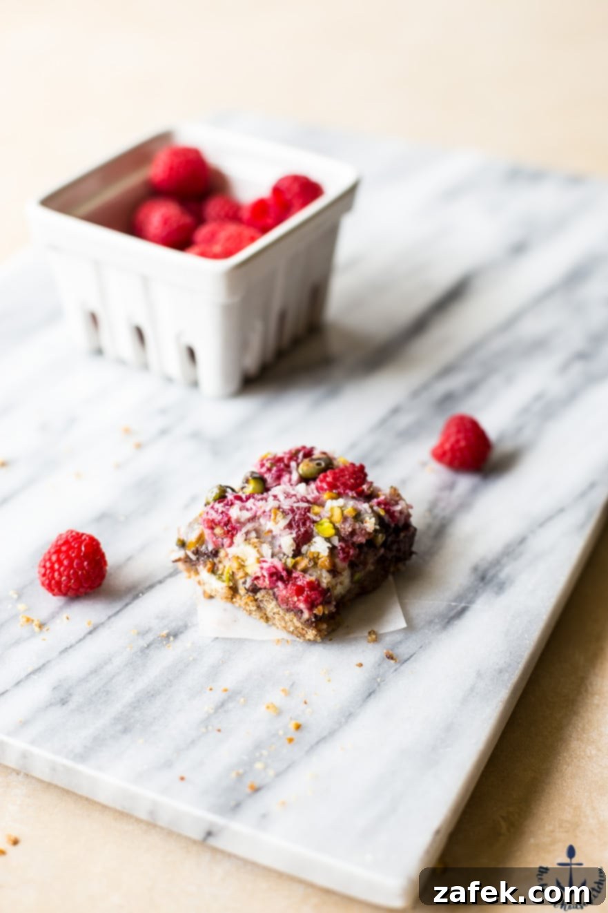 A single Raspberry Pistachio Magic Bar resting on a elegant marble board, with a container of fresh raspberries in the background, emphasizing its freshness and quality.
