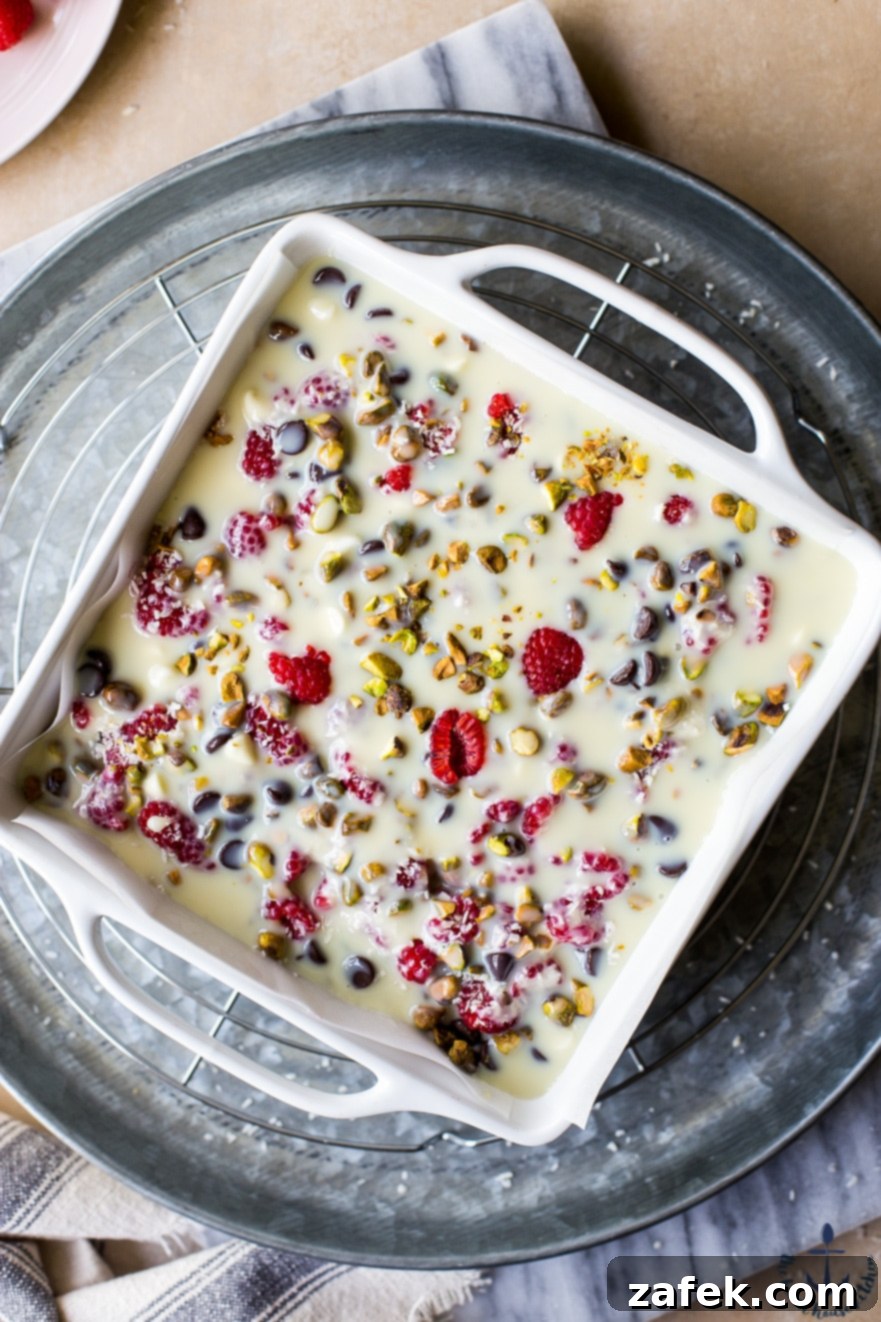 Overhead view of unbaked Raspberry Pistachio Magic Bars in a pan, with the colorful toppings distributed evenly over the pretzel crust, awaiting their transformation in the oven.
