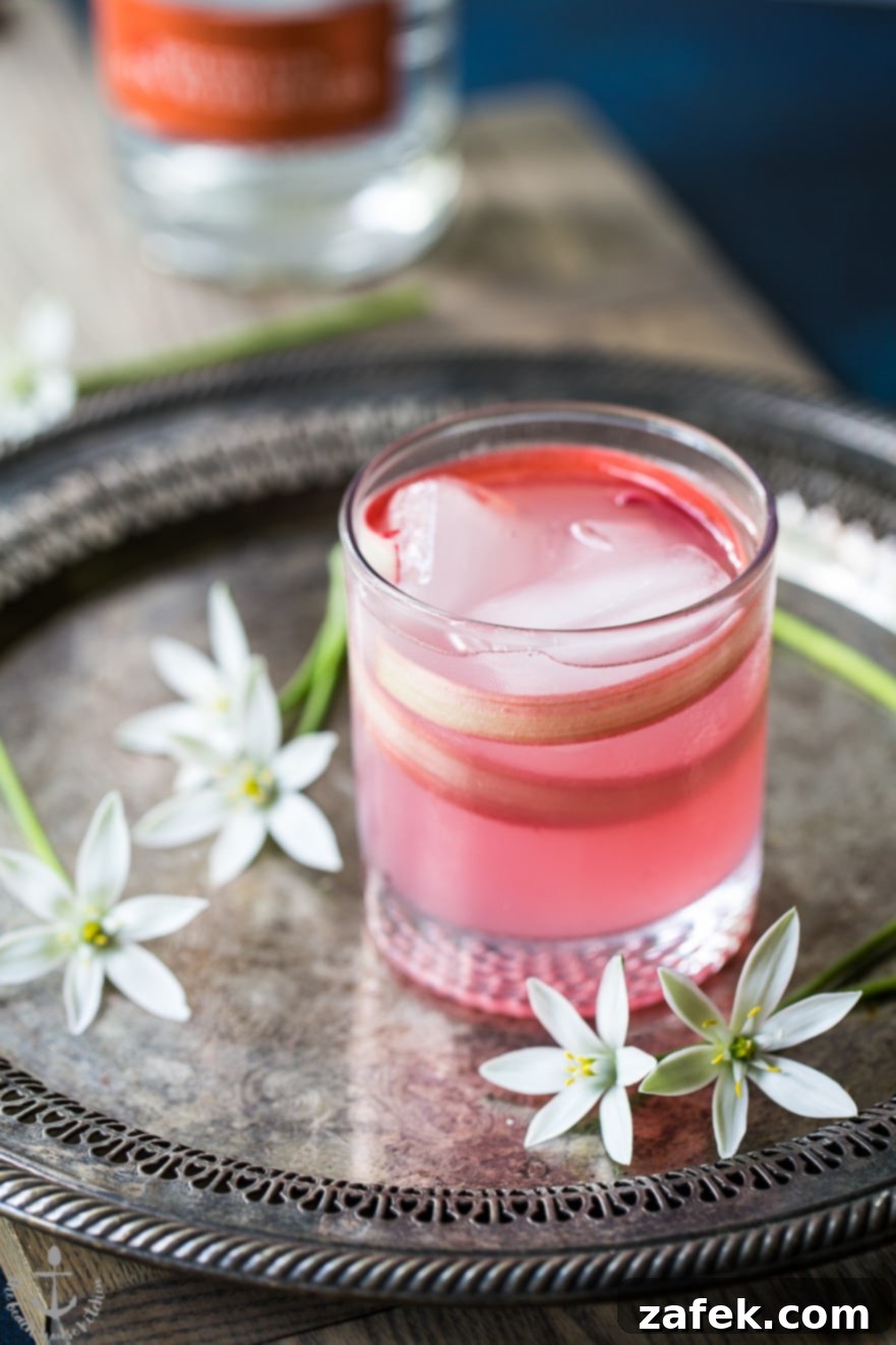 The Spiced Rhubarb Collins cocktail in a tall glass, showcasing its vibrant pink and clarity, presented on a silver tray with fresh flowers and citrus slices for an elegant touch.