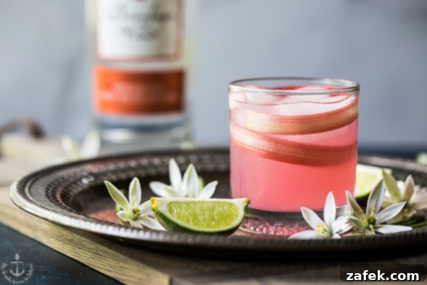 A refreshing Spiced Rhubarb Collins cocktail, garnished with a delicate rhubarb shaving, on a elegant silver tray surrounded by fresh limes and flowers, ready to serve.