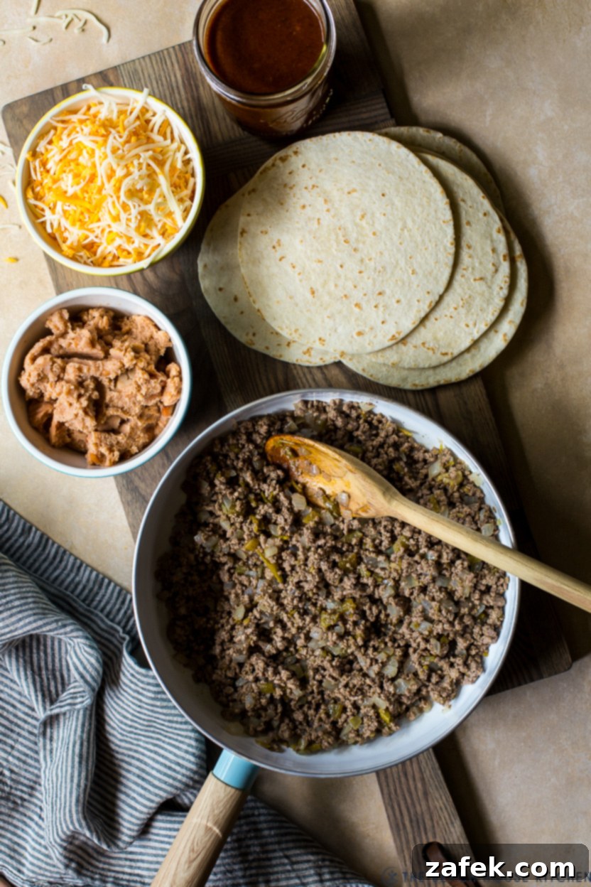 Effortless Homemade Beef Enchiladas 5 Overhead photo of a skillet of cooked ground beef, a bowl of shredded cheese and refired beans and flour tortillas