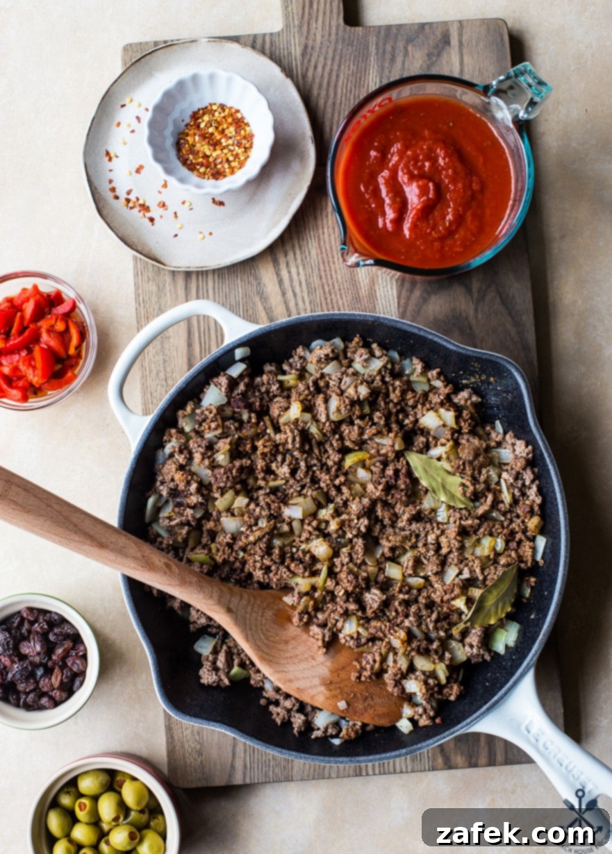 Close-up overhead photo of ground beef browning in a skillet with chopped onions and minced garlic, the aromatic base for picadillo