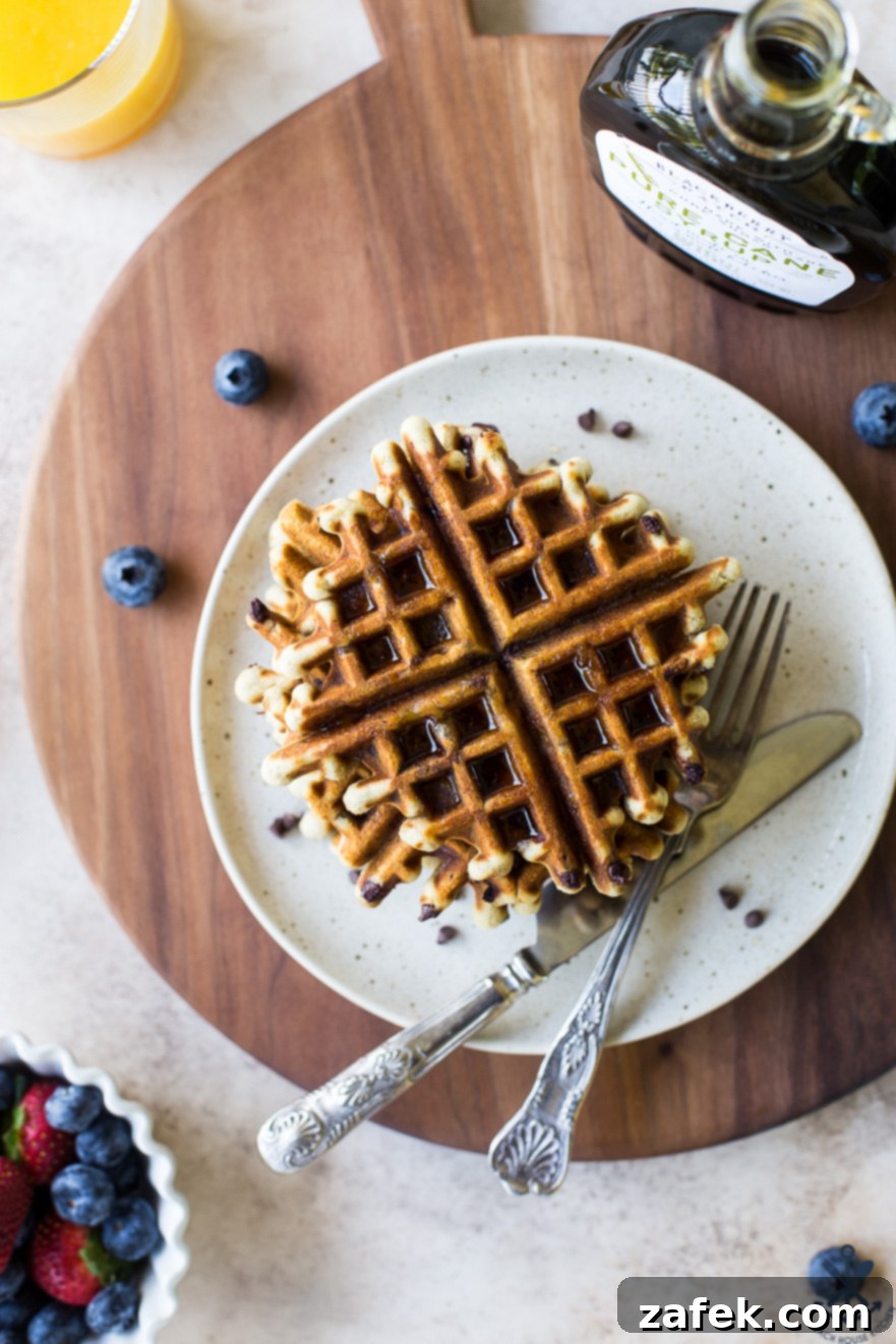 Ultimate Chocolate Chip Banana Bread Waffles 2 Overhead photo of golden-brown chocolate chip banana bread waffles on a white plate, resting on a rustic wooden board, garnished with fresh berries and a drizzle of maple syrup.