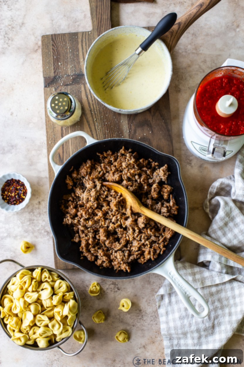 Overhead photo of a skillet of cooked crumbled Italian sausage and a bowl of tortellini and a saucepan of alfredo sauce